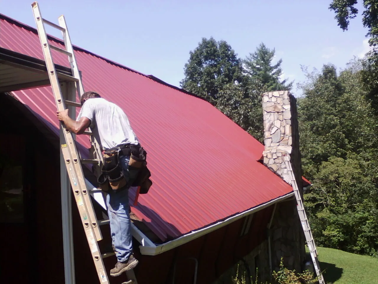 Roofer on a ladder beside a red metal roof and stone chimney — Soffit & Fascia Repair by Roofing Brothers in Scammon